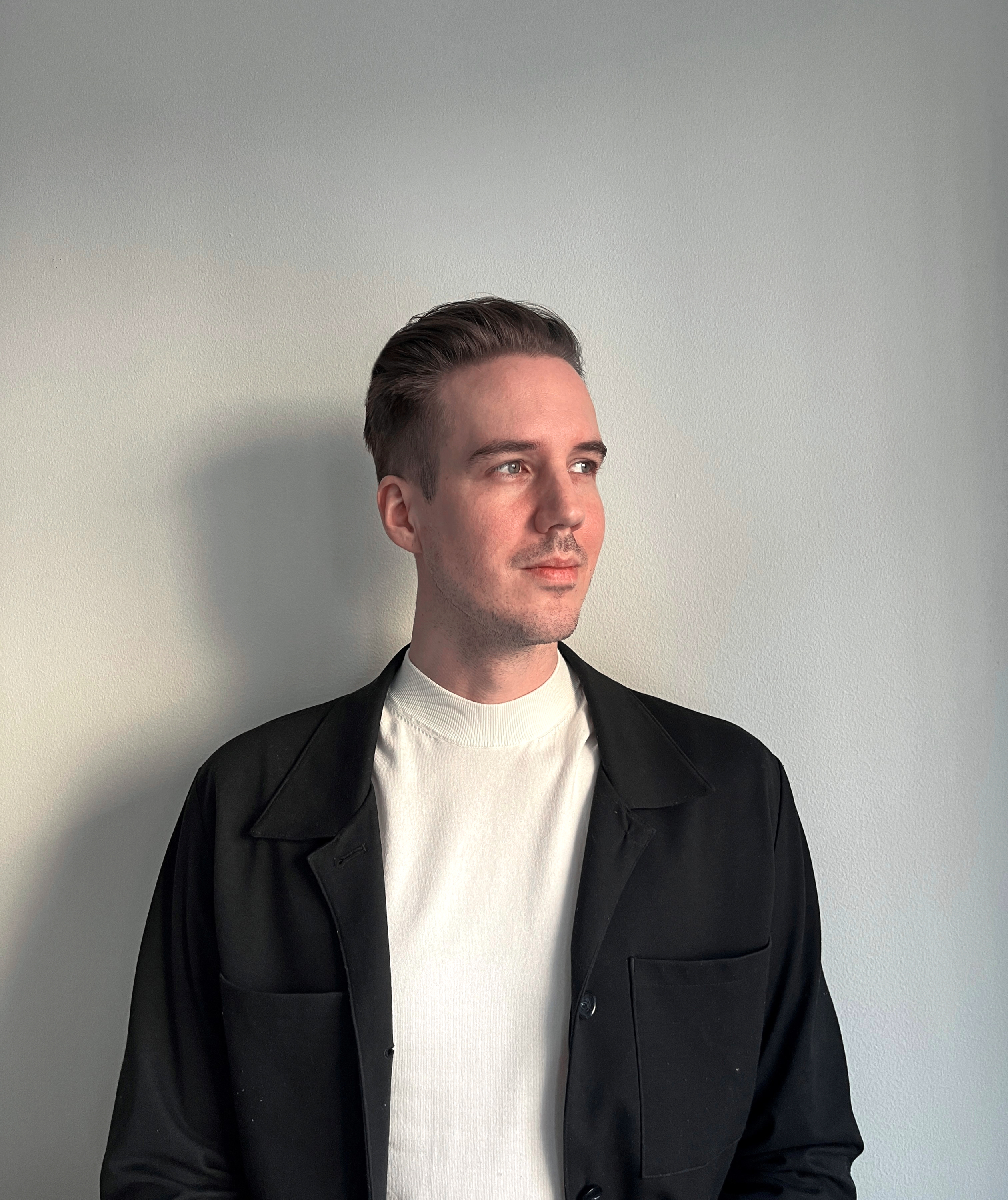 En man med kort hår, vit skjorta och svart jacka står mot en ljus vägg och tittar åt höger.