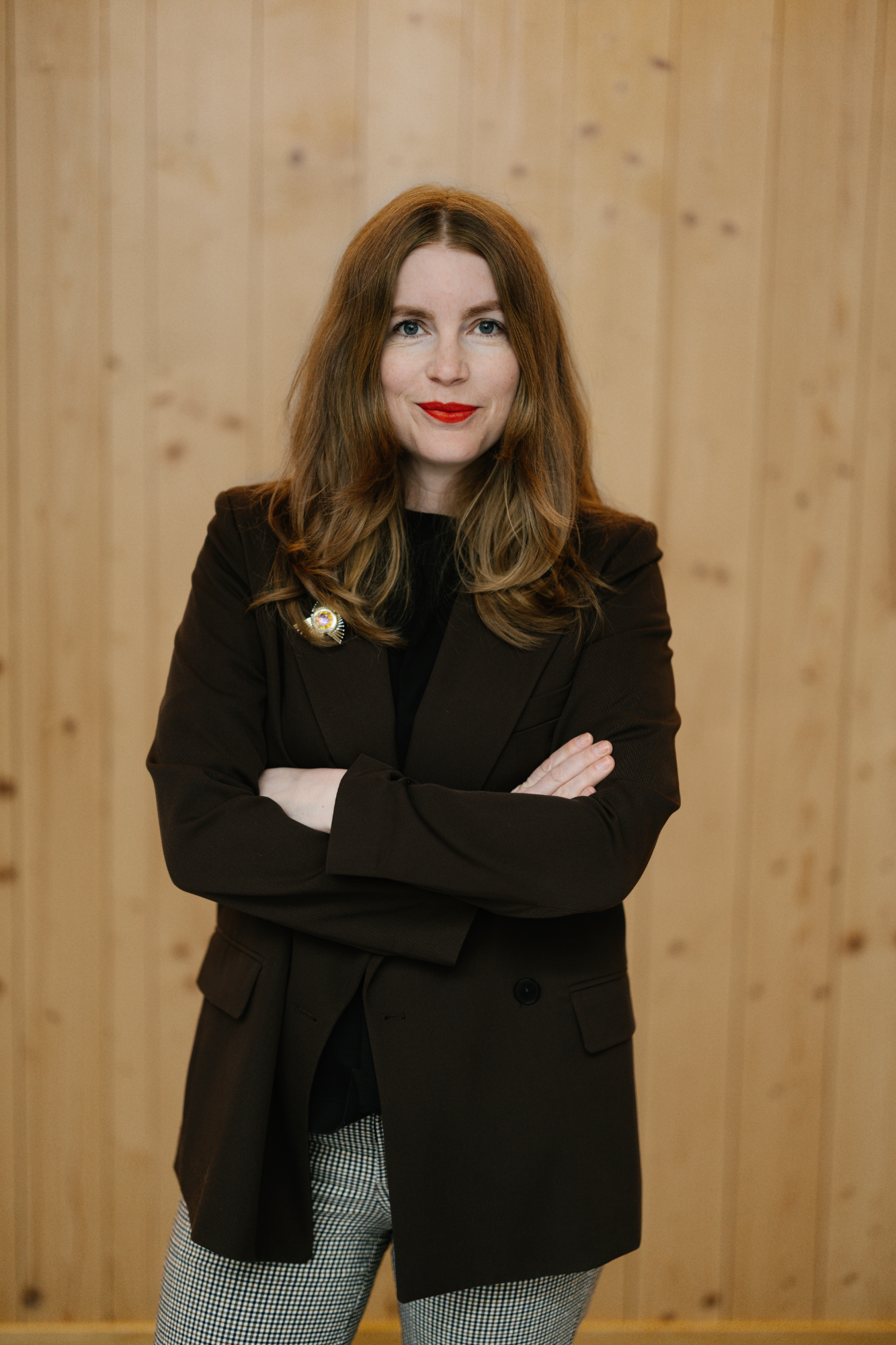 En kvinna med långt brunt hår, rött läppstift och en mörk kavaj står med korslagda armar framför en ljus trävägg.