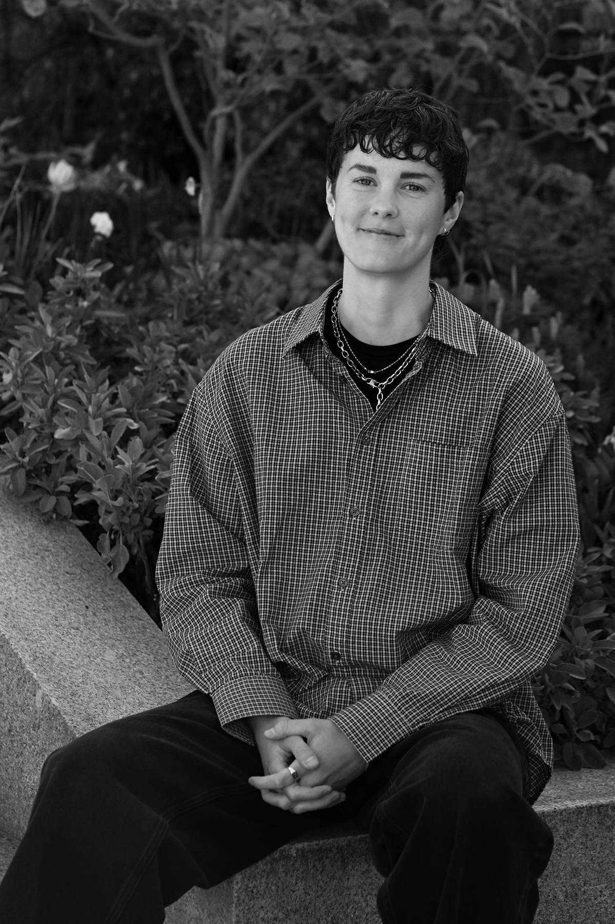 En person med kort mörkt hår, klädd i en rutig skjorta och halsband, sitter på en bänk av sten utomhus med växter i bakgrunden.