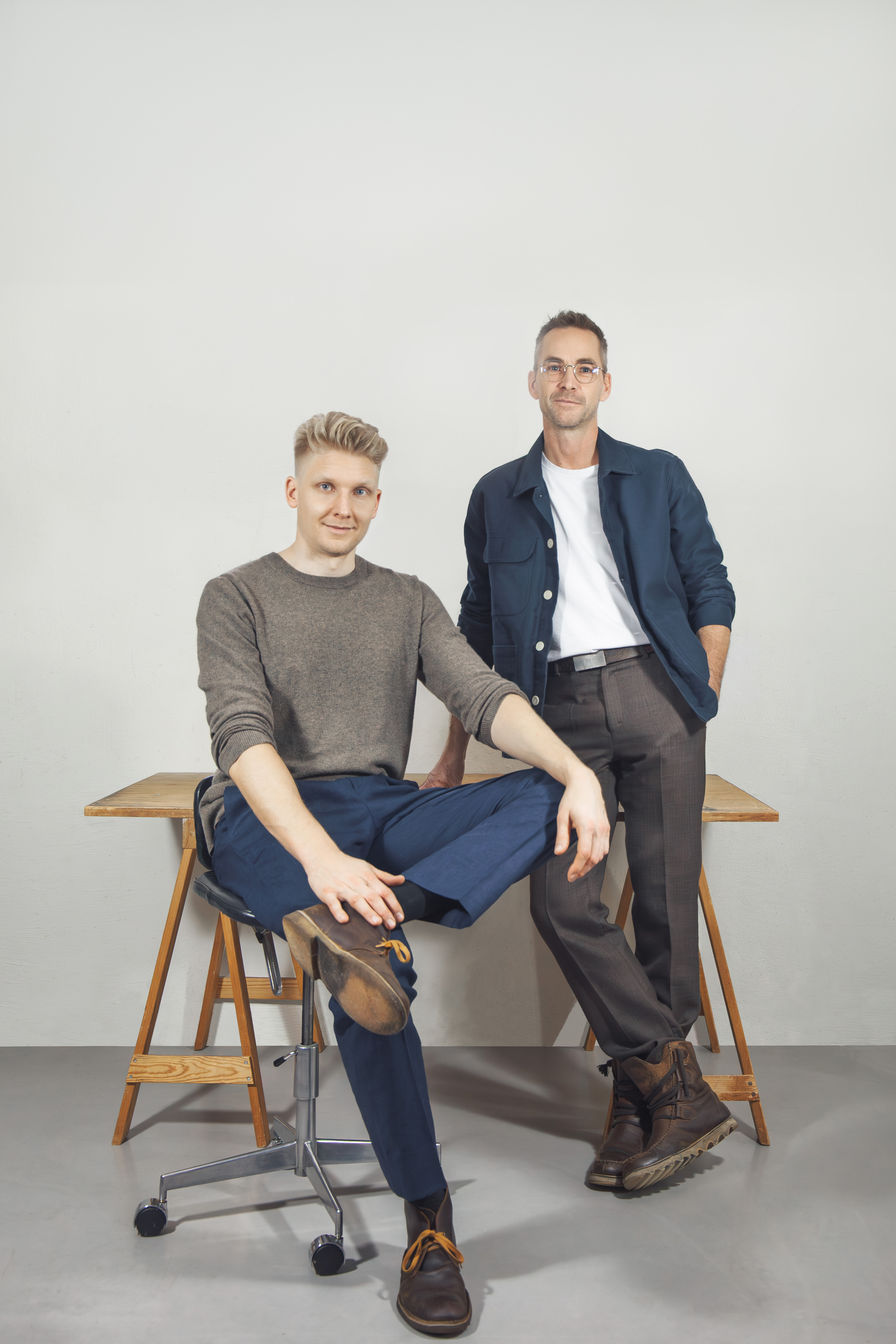 Två män poserar i ett kontor; en sitter på en stol med benen i kors och den andra står bredvid, båda tittar mot kameran.