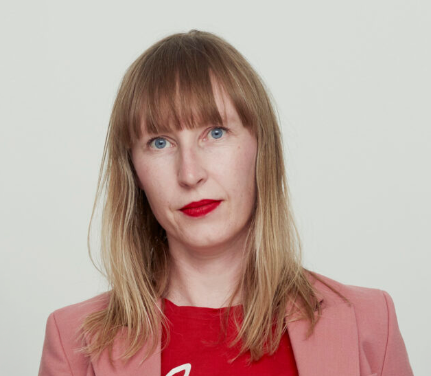 En kvinna med rakt blont hår och lugg, har rött läppstift, röd topp och rosa kavaj, står framför en ljus bakgrund.