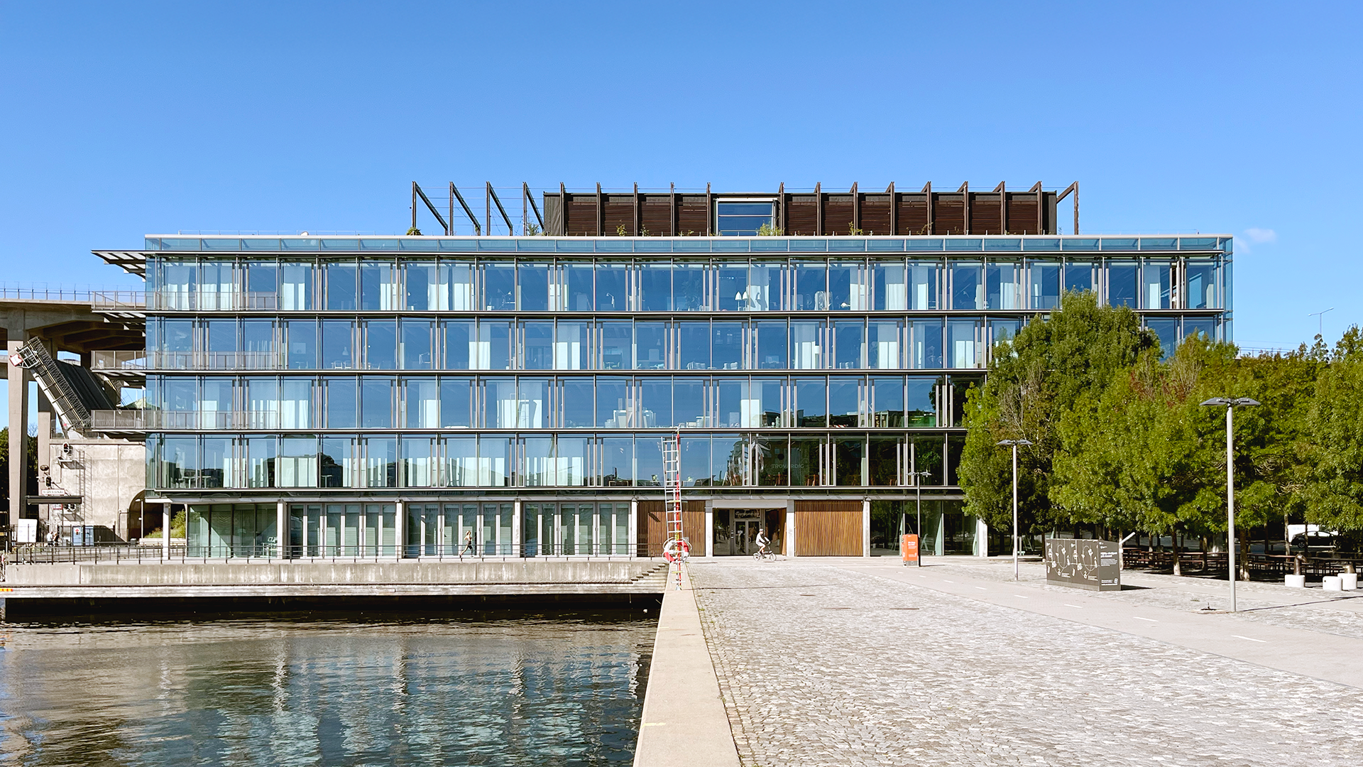 En byggnad med glasfasad står vid vattnet, med träd till höger och en klar blå himmel ovanför.