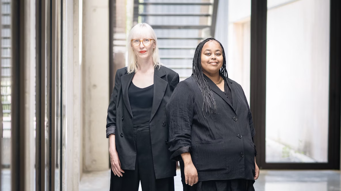 Två kvinnor i svarta kläder står bredvid varandra i en hall med stora fönster och väggar av betong.