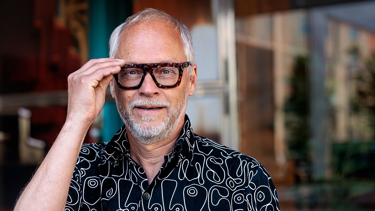 En man med grått hår och skägg står utomhus. Han har mönstrad skjorta och stora glasögon, håller i glasögonen och ler mot kameran.