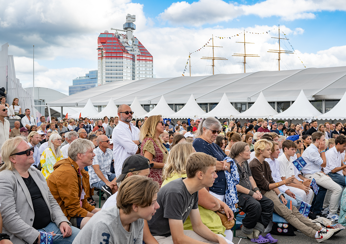 En folkmassa sitter och står utomhus vid ett evenemang. I bakgrunden finns vita tält, ett segelfartyg och en byggnad.