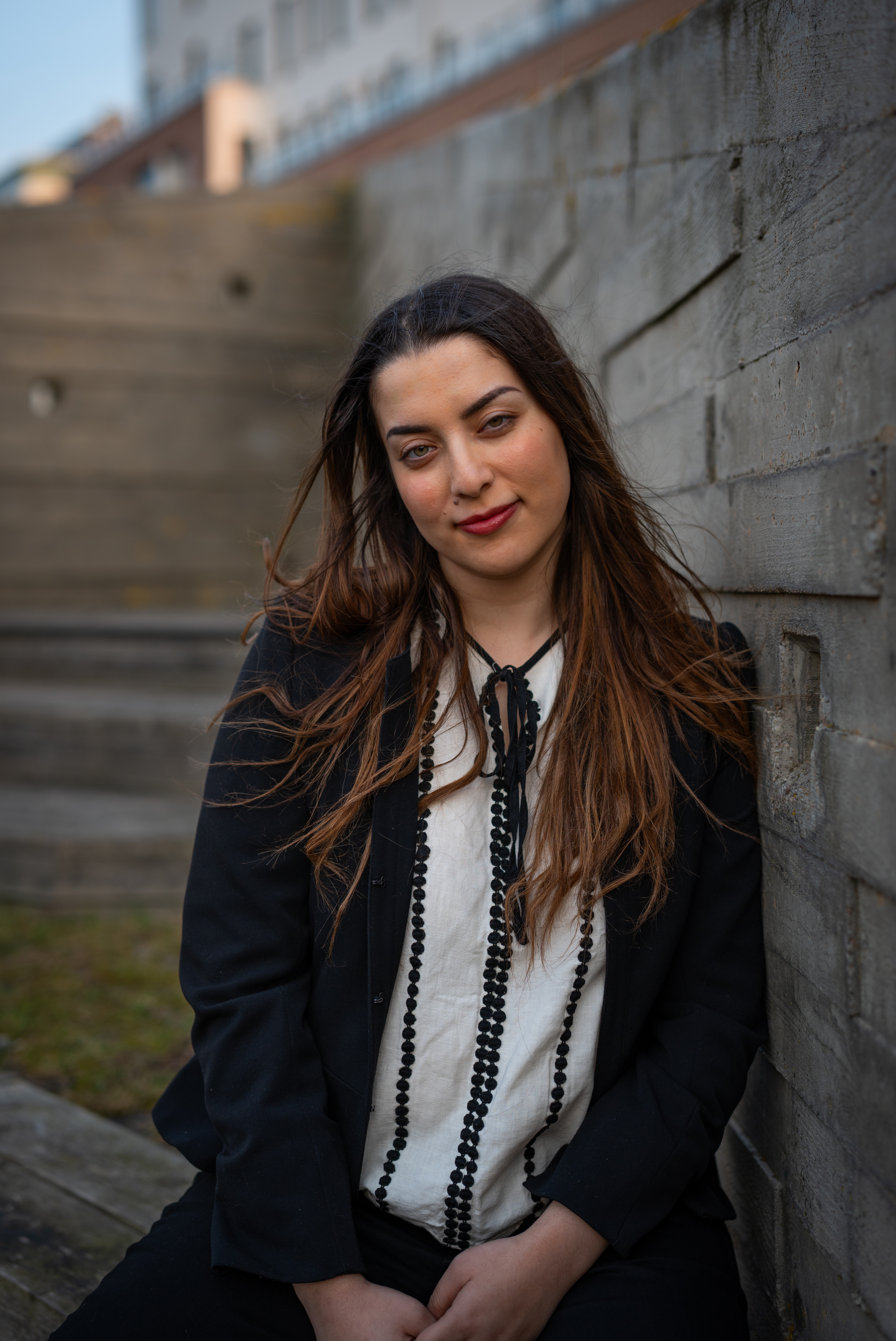 En kvinna med långt brunt hår, klädd i svart kavaj och vit blus, sitter utomhus mot en betongvägg och tittar mot kameran.
