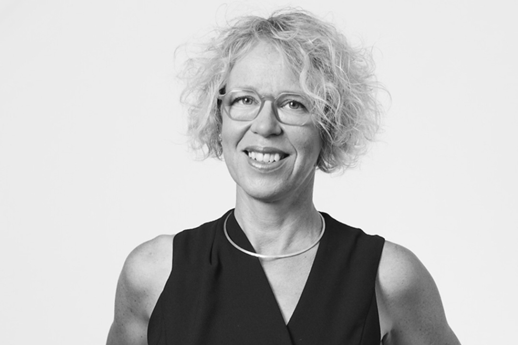 En kvinna med kort, lockigt blont hår, glasögon, ärmlös mörk topp och halsband ler mot kameran framför en enkel bakgrund.