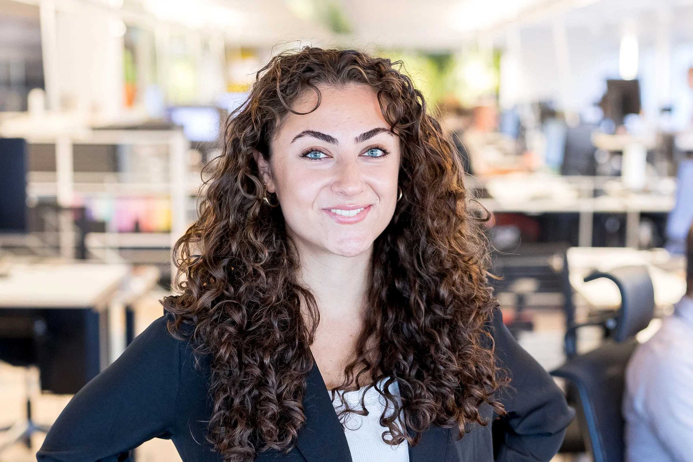 En kvinna med långt lockigt hår och svart kavaj står i ett kontor och ler mot kameran.