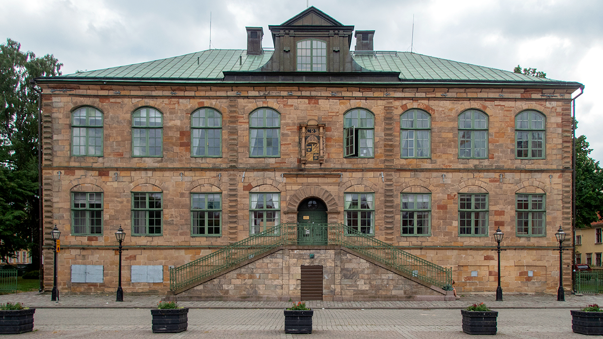 En rektangulär stenbyggnad med många fönster, ett grönt plåttak och en mittentrappa som leder till huvudingången.