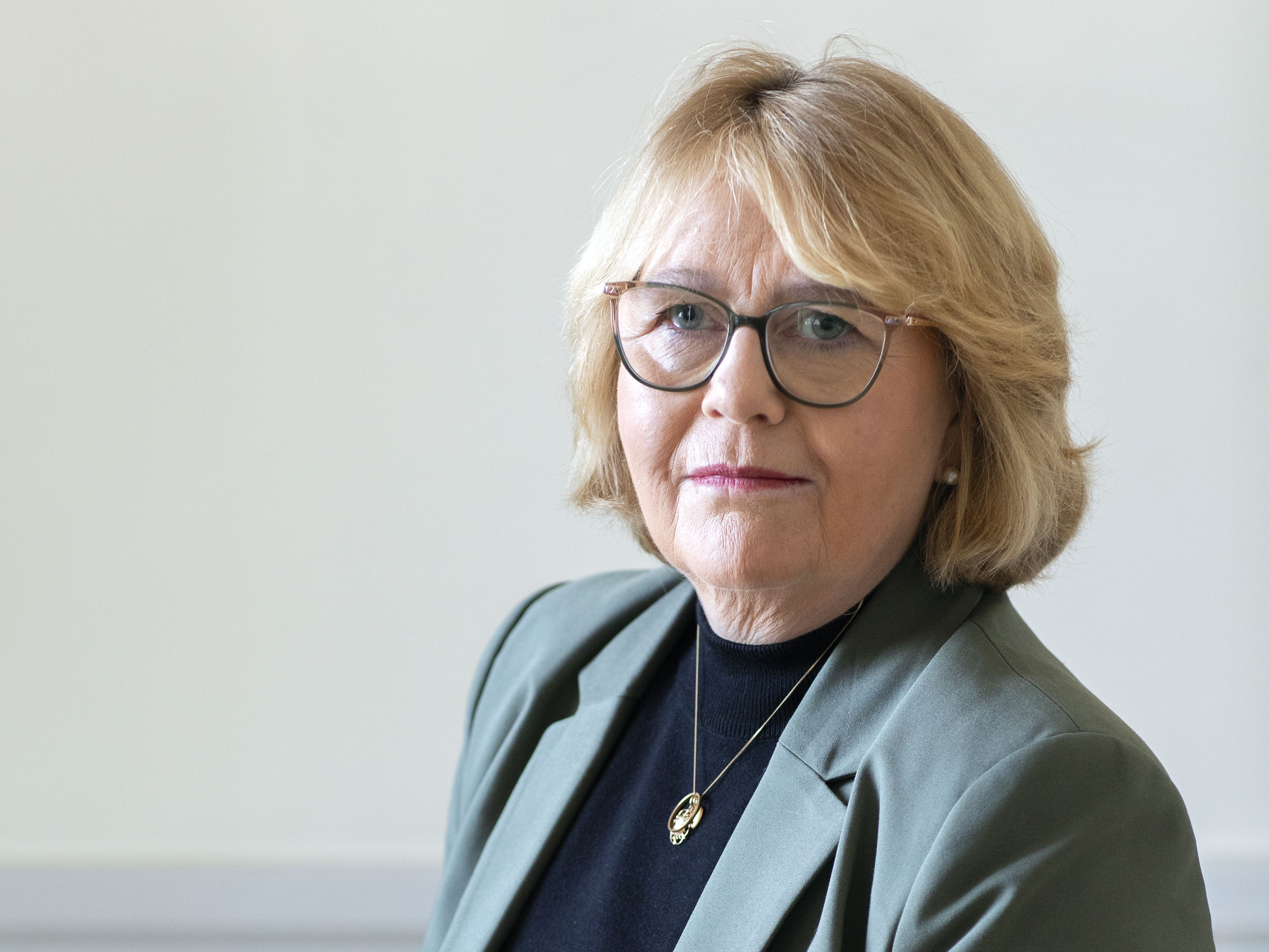 En kvinna med kort blont hår och glasögon har på sig en grön kavaj och svart topp, tittar mot kameran mot ljus bakgrund.