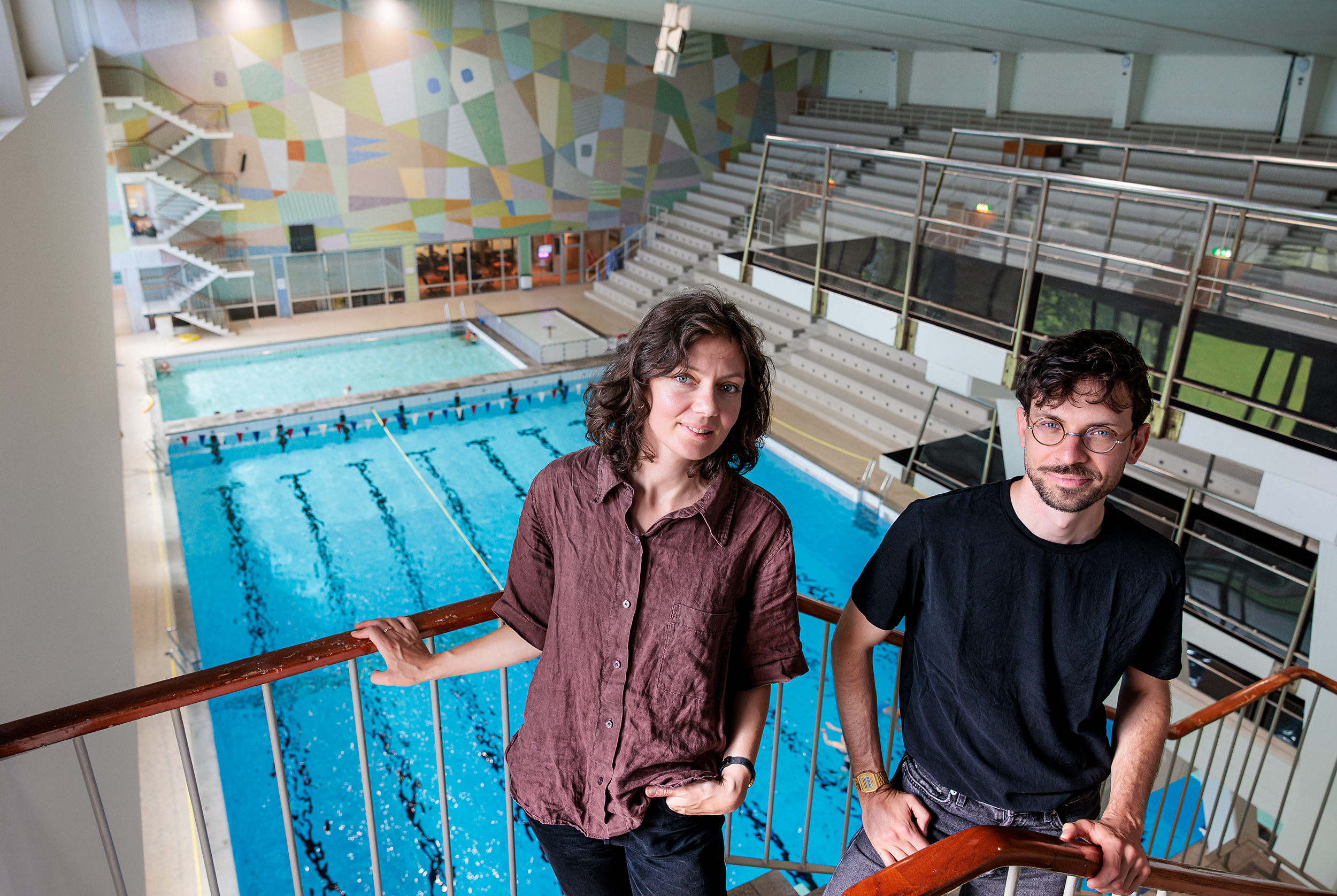 Två personer står på en inomhusbalkong och ser ut över en tom simbassäng med banmarkeringar och läktare.