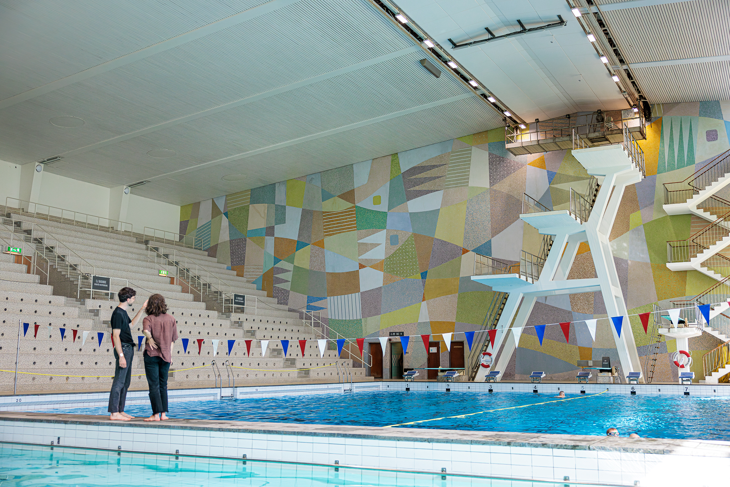 Två personer står bredvid en inomhuspool med ett högt hopptorn och geometrisk väggkonst i bakgrunden.