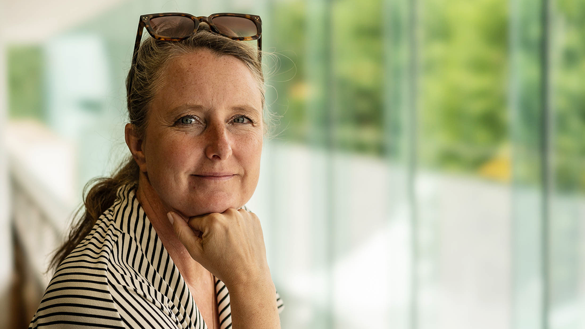 En kvinna med solglasögon på huvudet och randig tröja vilar hakan i handen och tittar mot kameran. Fönster och växter syns bakom.