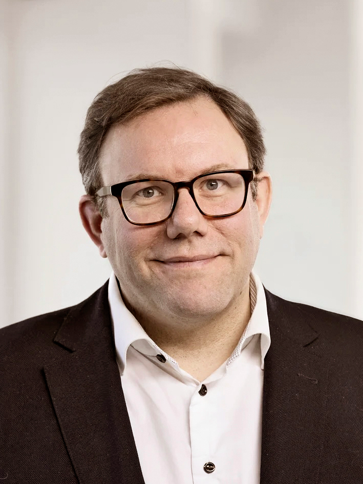 En man med kort brunt hår och glasögon står i mörk kavaj och vit skjorta framför en ljus, suddig bakgrund.