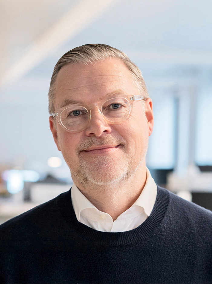 En man med glasögon och kort skägg, vit skjorta och mörk tröja står i ett kontor.