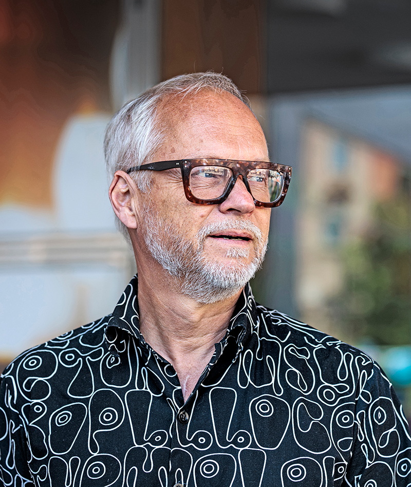 En man med vitt hår, skägg och stora glasögon står utomhus och har på sig en svart skjorta med vitt mönster.