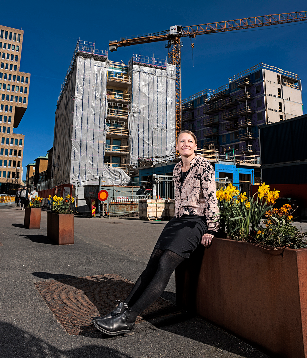 En person sitter på en planteringslåda med gula blommor i ett urbant område, med byggarbetsplatser, kranar och byggnader i bakgrunden.