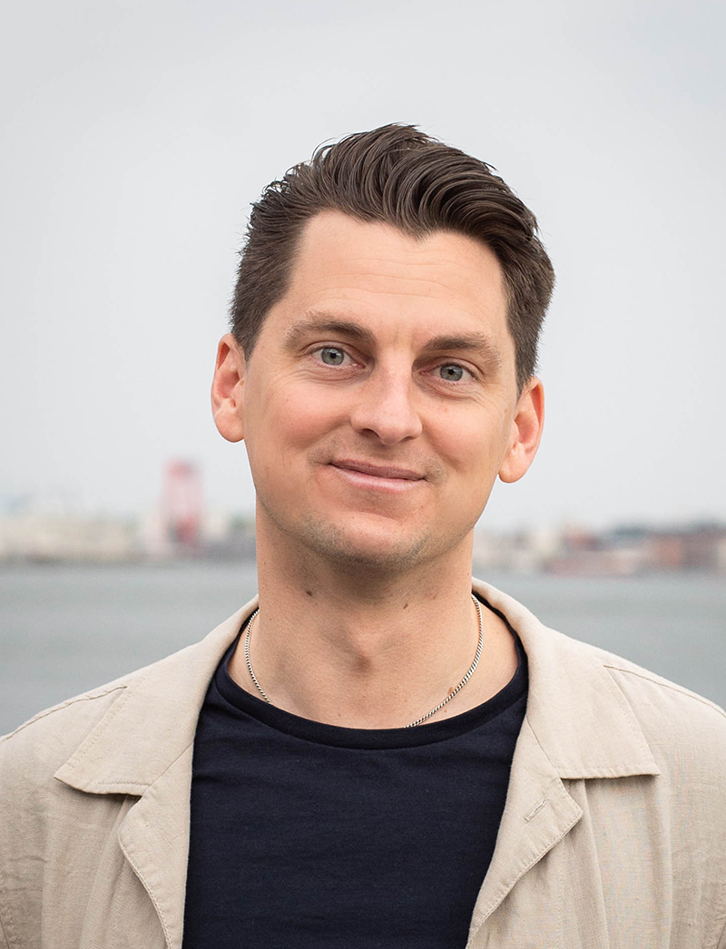 En man med kort brunt hår och beige jacka över en mörk skjorta står utomhus vid vatten med byggnader i bakgrunden.