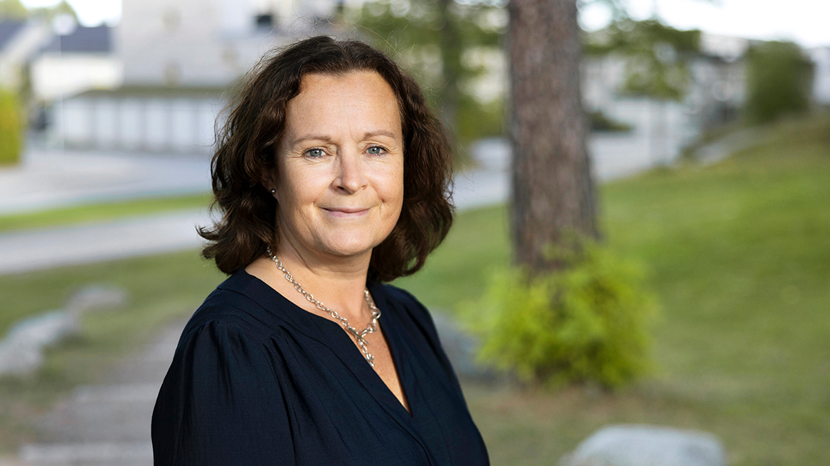 En kvinna med brunt axellångt hår och en marinblå blus står utomhus och ler svagt, med gräs och byggnader i bakgrunden.