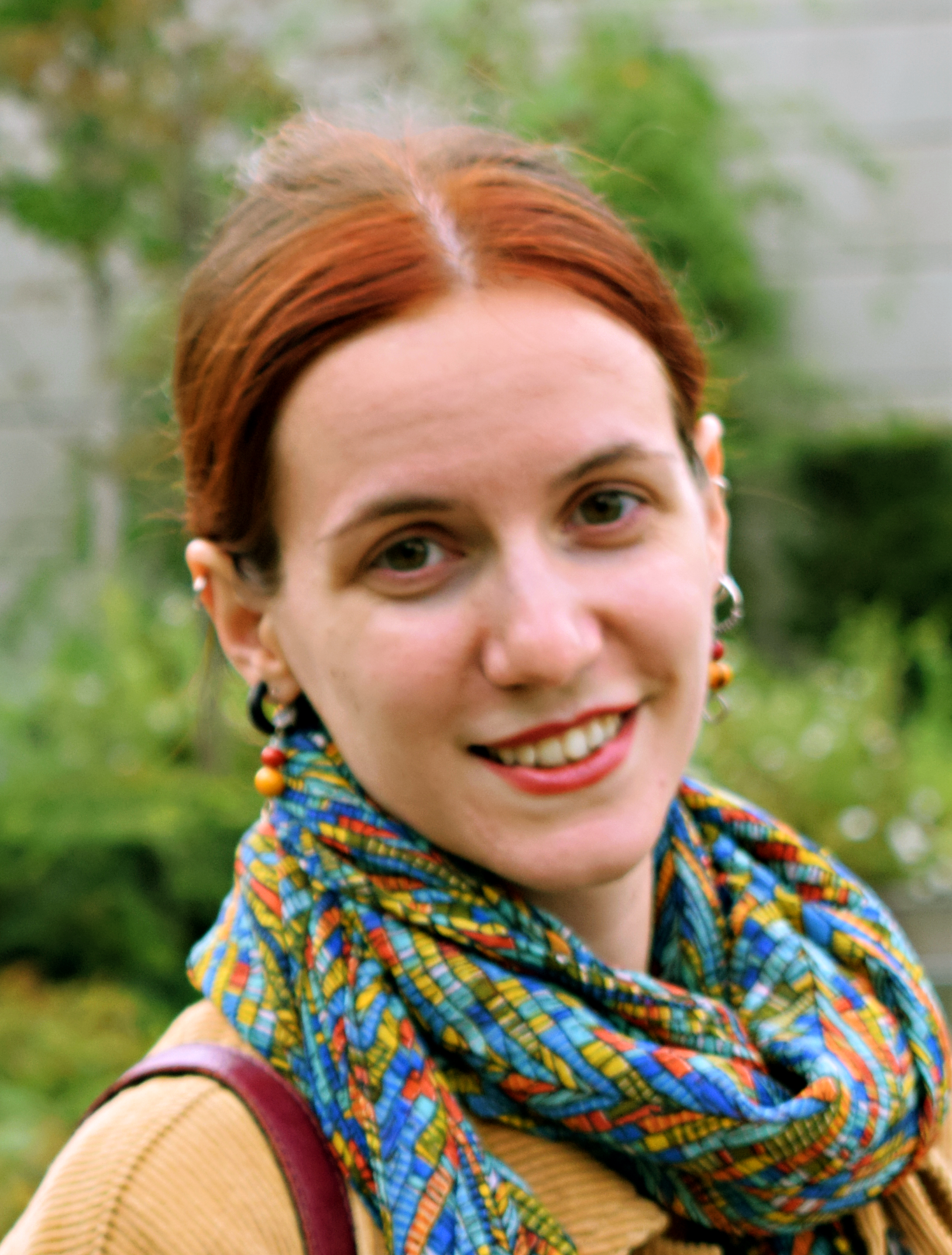 En kvinna med rött hår, som har en scarf och örhängen på sig, ler utomhus med växtlighet i bakgrunden.