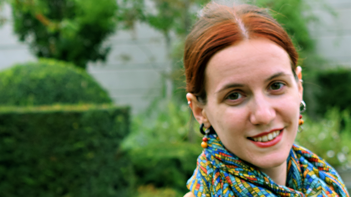 En kvinna med rött hår och en mönstrad scarf ler utomhus, med gröna växter och klippta buskar i bakgrunden.