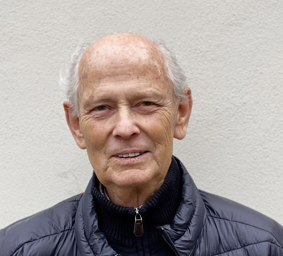 En äldre man med tunt vitt hår och svart jacka står framför en ljus vägg och tittar mot kameran.