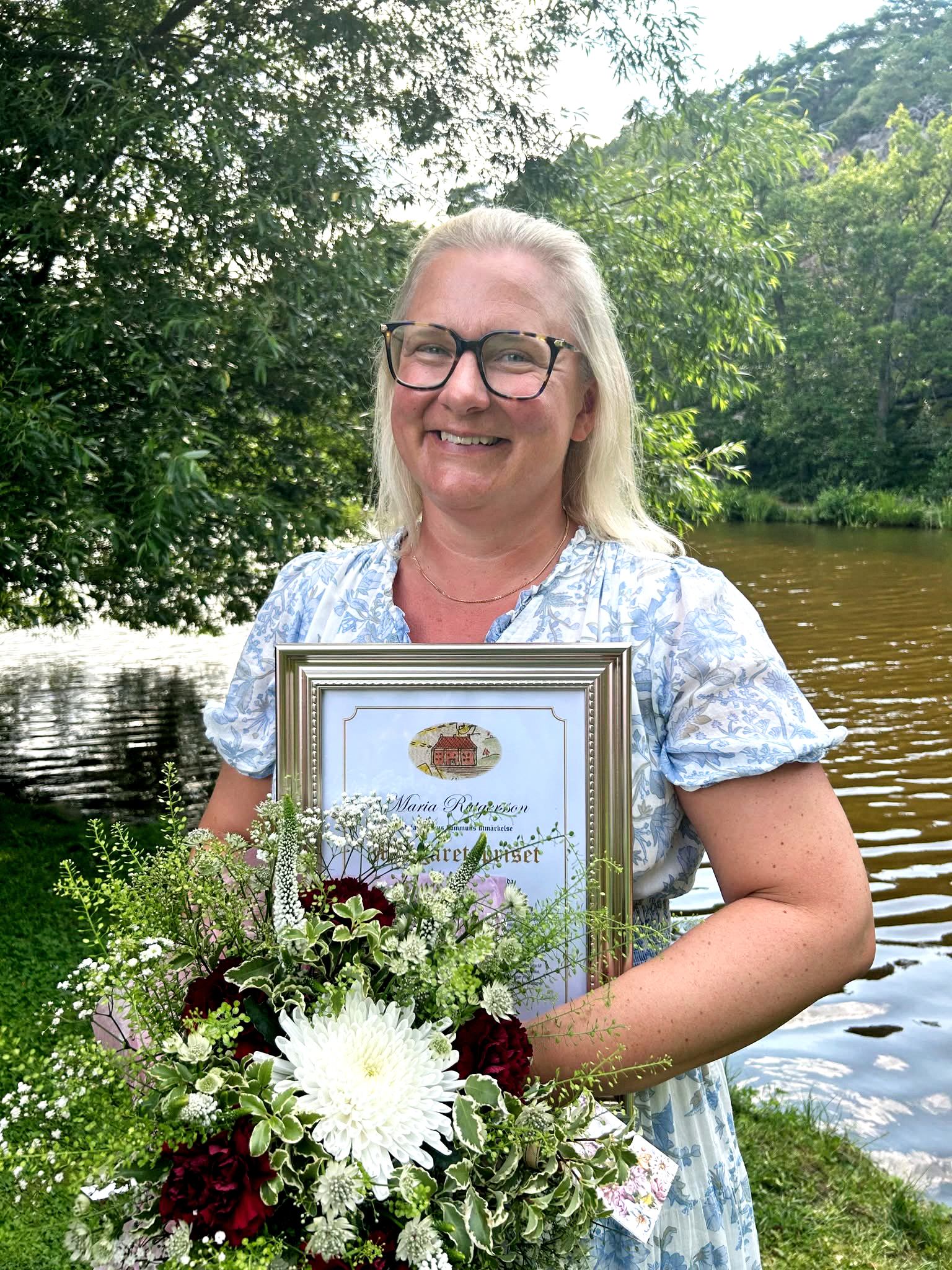 En kvinna med glasögon står utomhus vid en damm och håller ett inramat diplom och en blombukett.