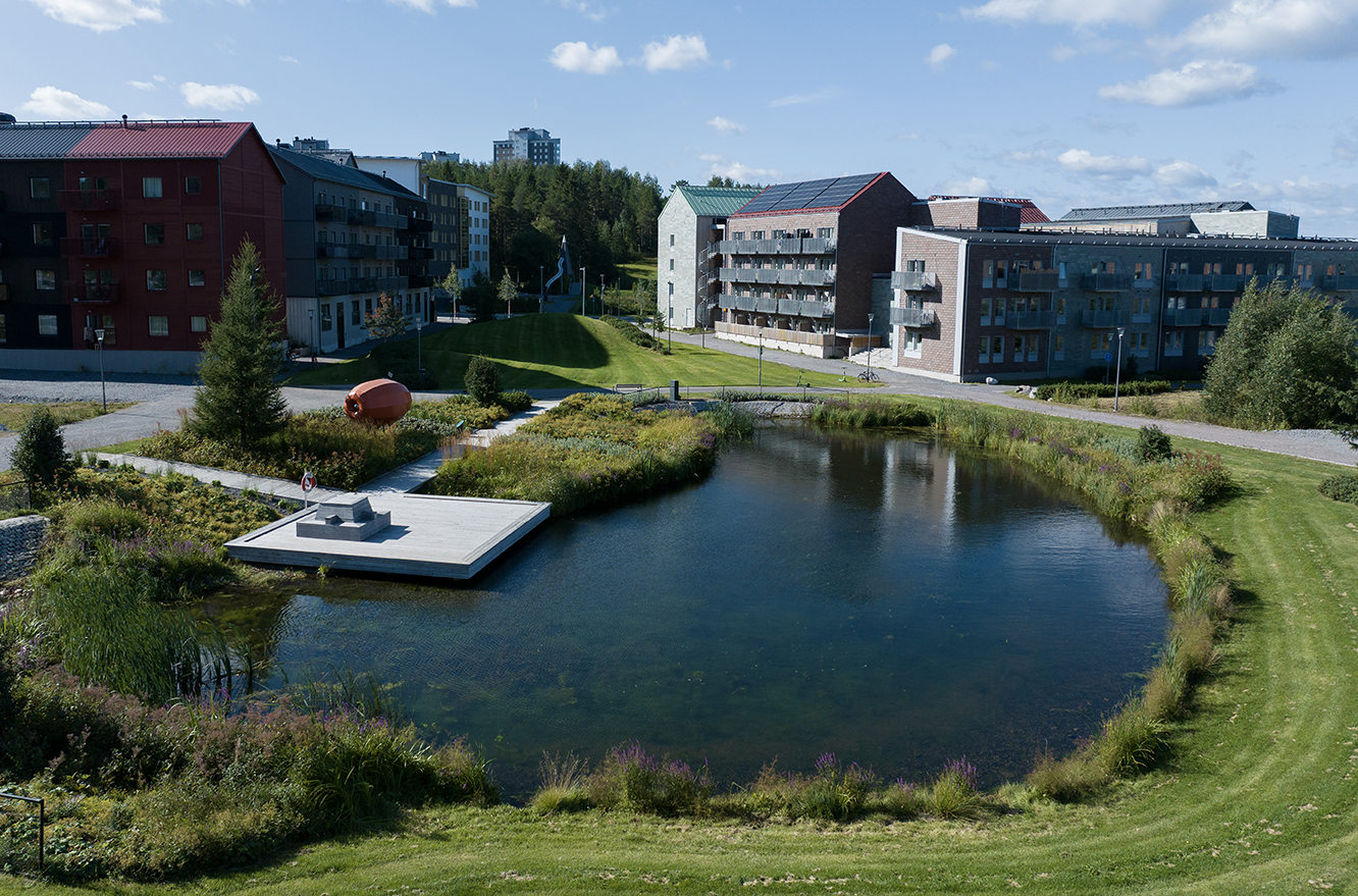 En liten damm med en brygga omges av gräs, växter och moderna lägenhetshus under en blå himmel med moln.
