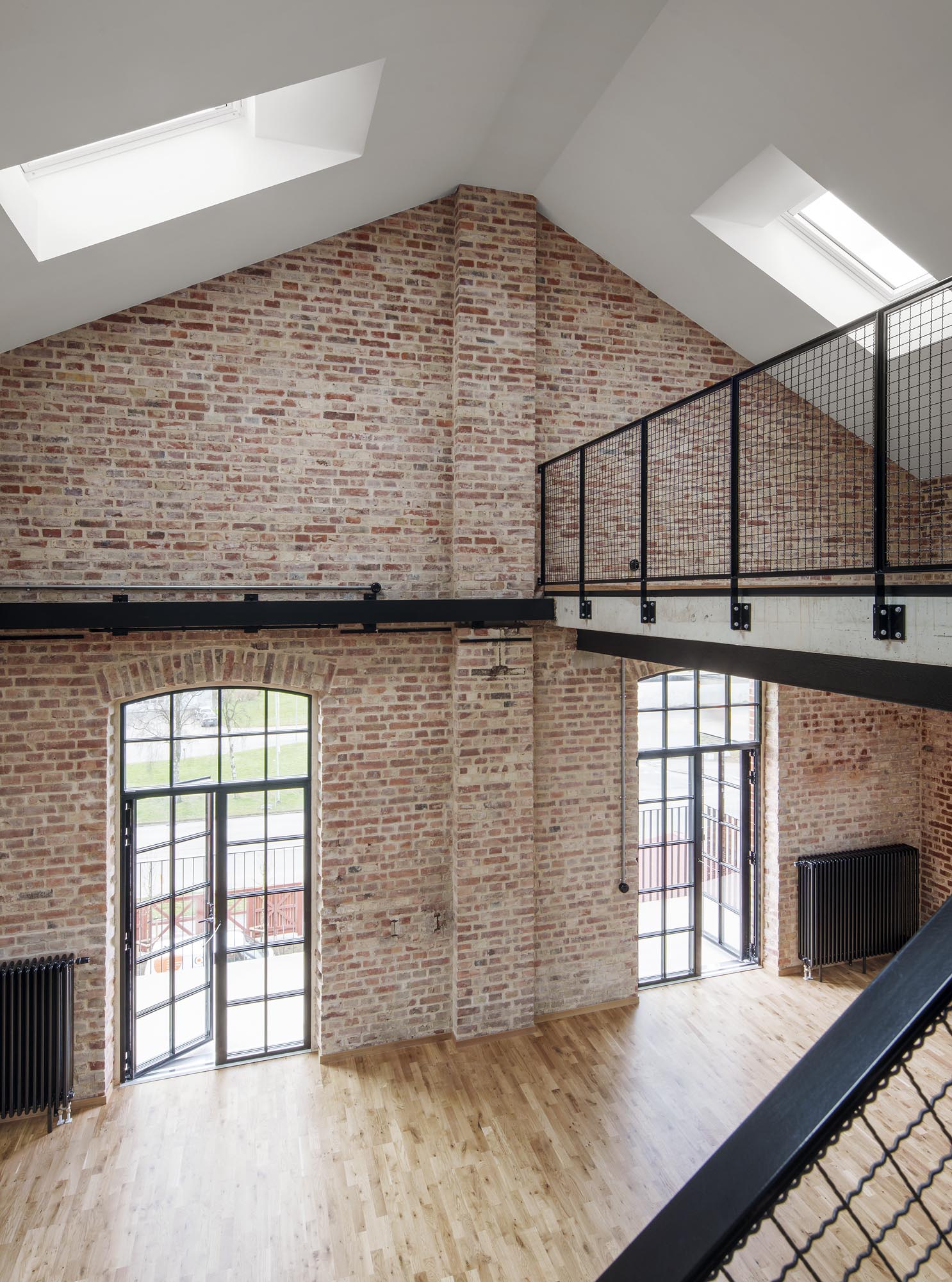 Loft med synliga tegelväggar, stora välvda fönster, svarta metallräcken, takfönster och trägolv samt dagsljus i rummet.