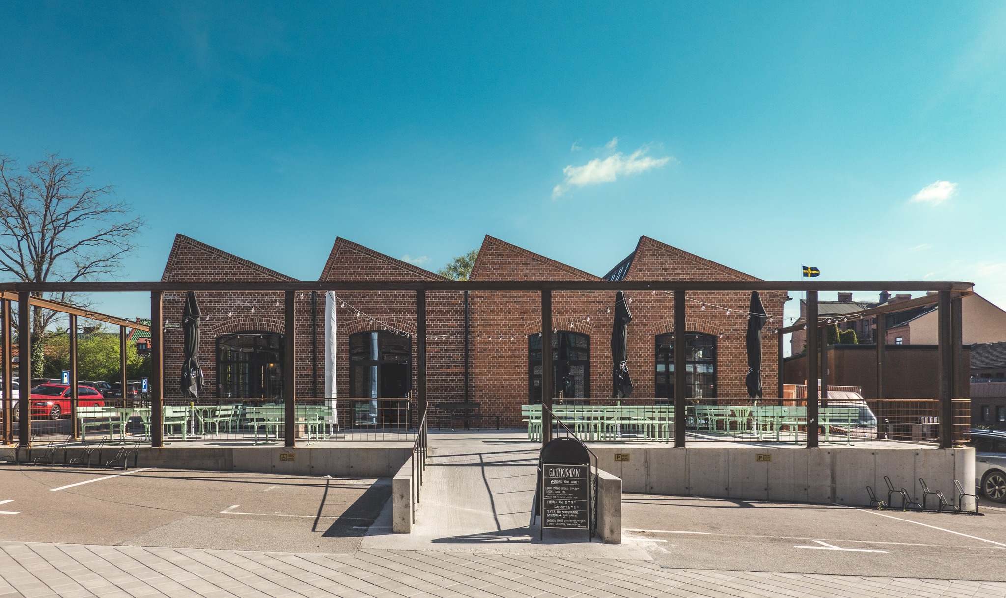 Utomhus sittplatser framför en tegelbyggnad med vinklade tak, ljusslingor och tomma bord under dagsljus.