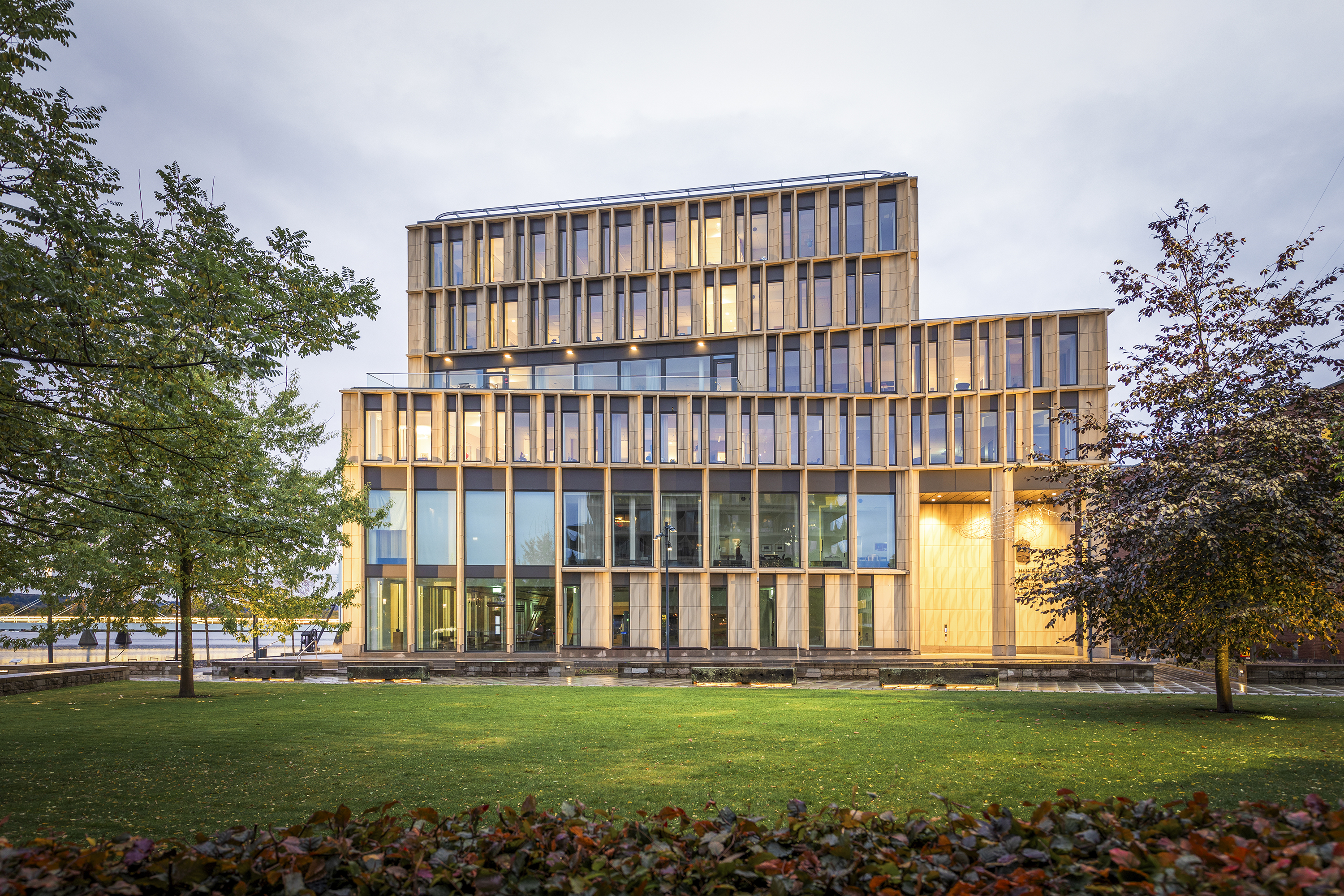 Flerårig byggnad med stora glasfönster och vertikala träpaneler, omgiven av gräsmatta och träd under molnig himmel.