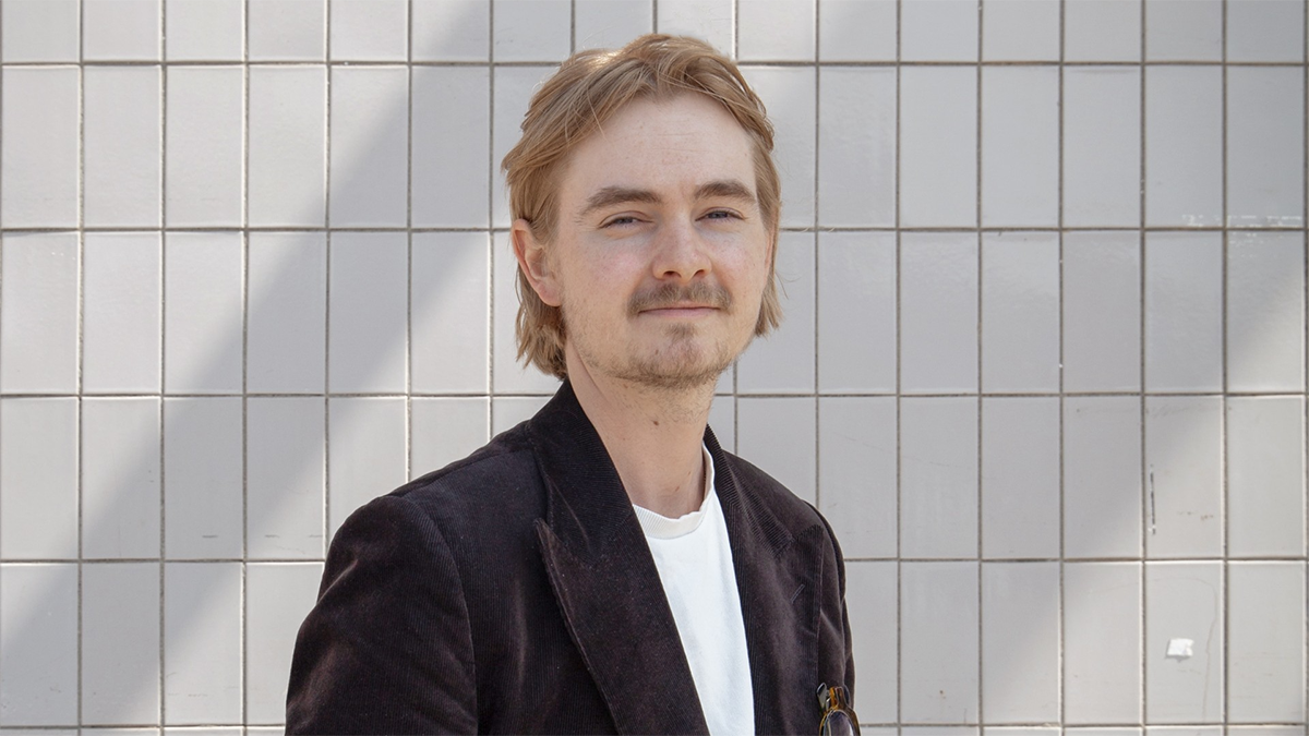 En person med ljust brunt hår och mustasch står framför en kaklad vägg och har på sig en mörk kavaj över en vit skjorta.