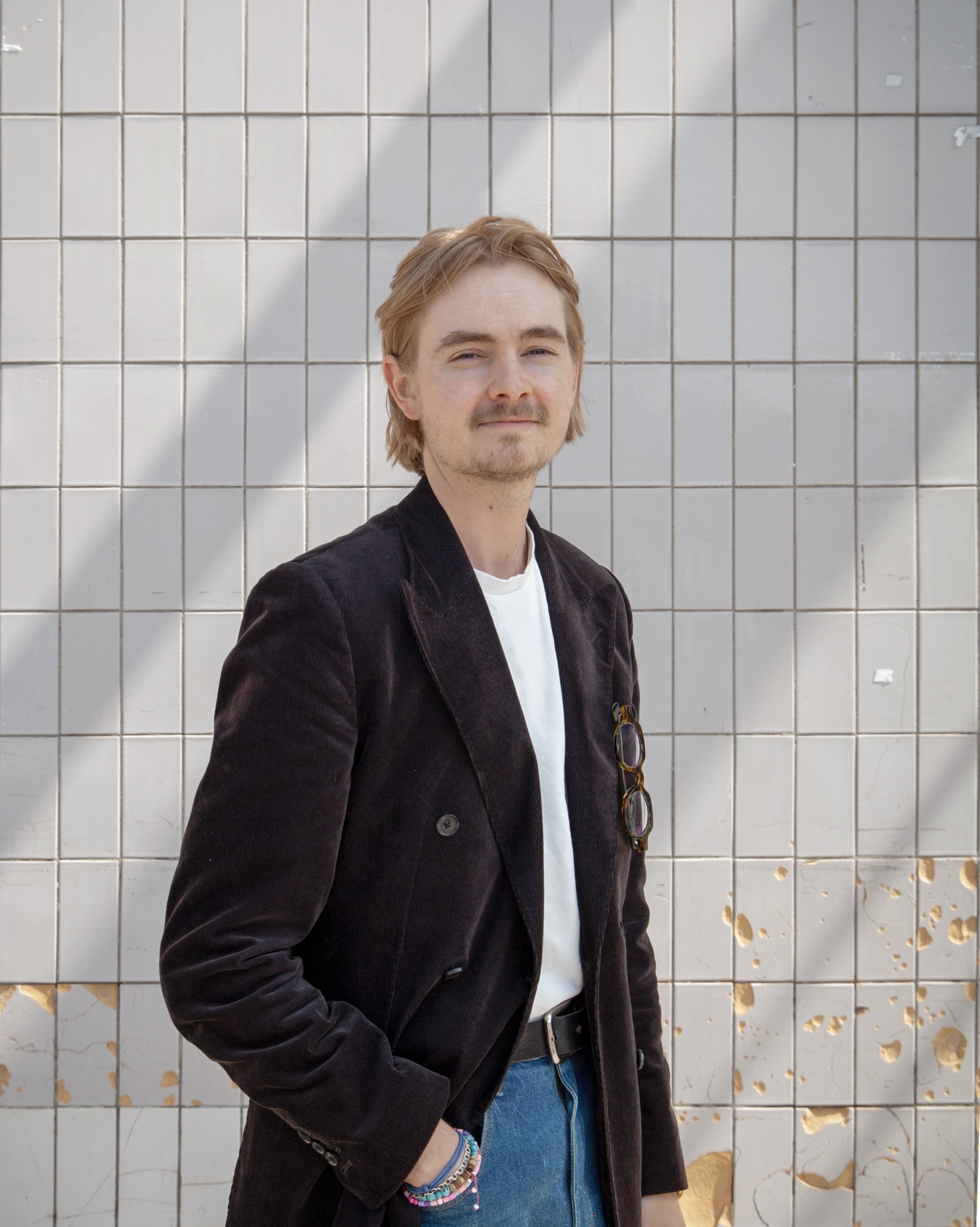 En person med ljusbrunt hår och mustasch står framför en kaklad vägg, iförd kavaj, vit skjorta och blå jeans, med en hand i fickan.