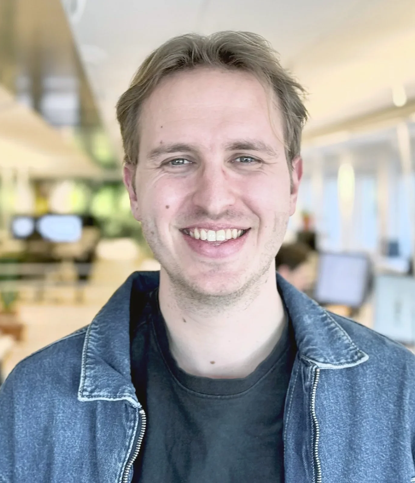 En man med ljust brunt hår, iklädd blå jacka över svart tröja, ler mot kameran i ett kontorsutrymme.