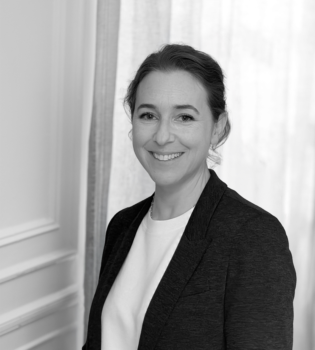 En kvinna med kavaj och ljus topp står inomhus, ler mot kameran. Bakom henne finns en gardin och väggpanel. Bilden är svartvit.