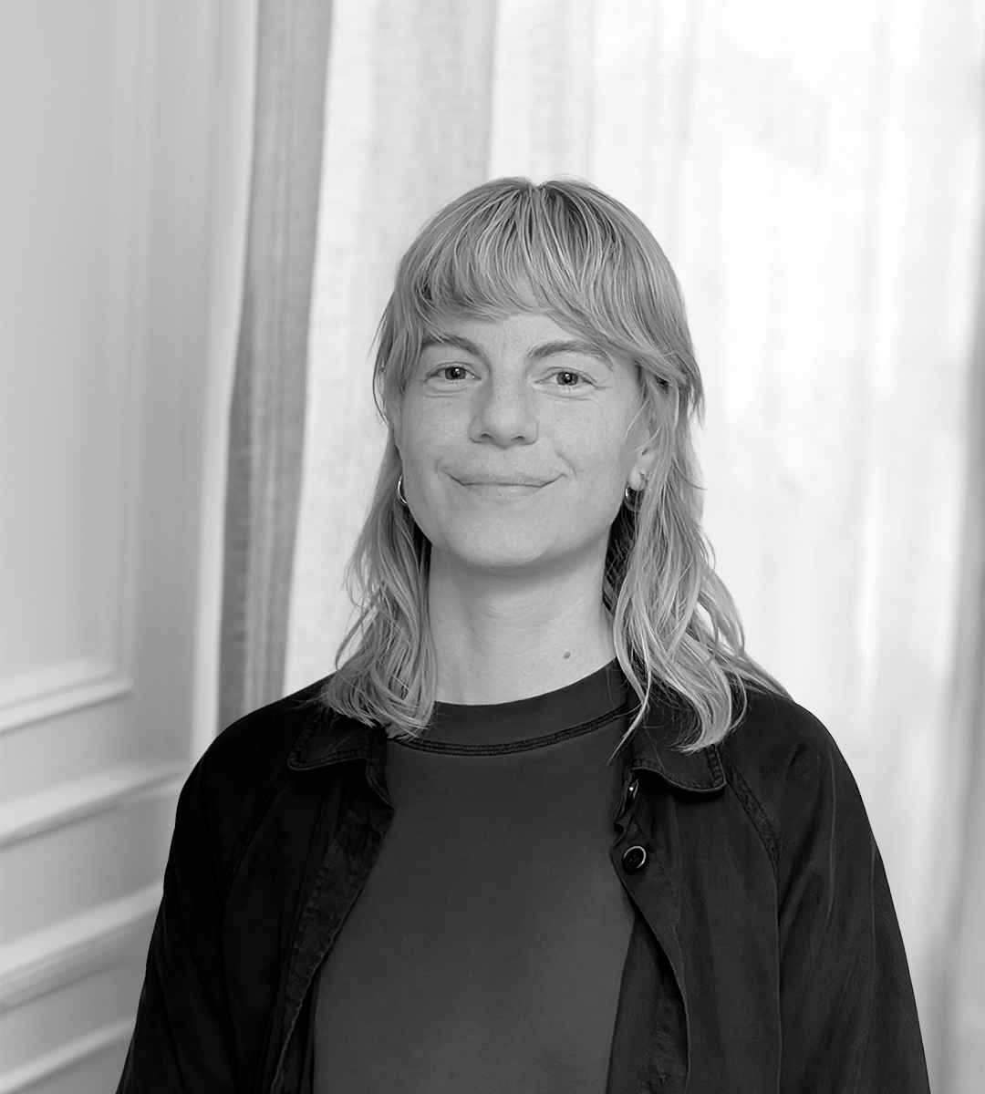 En person med ljust axellångt hår i mörk tröja och jacka står inomhus framför ett fönster med gardiner och tittar mot kameran.