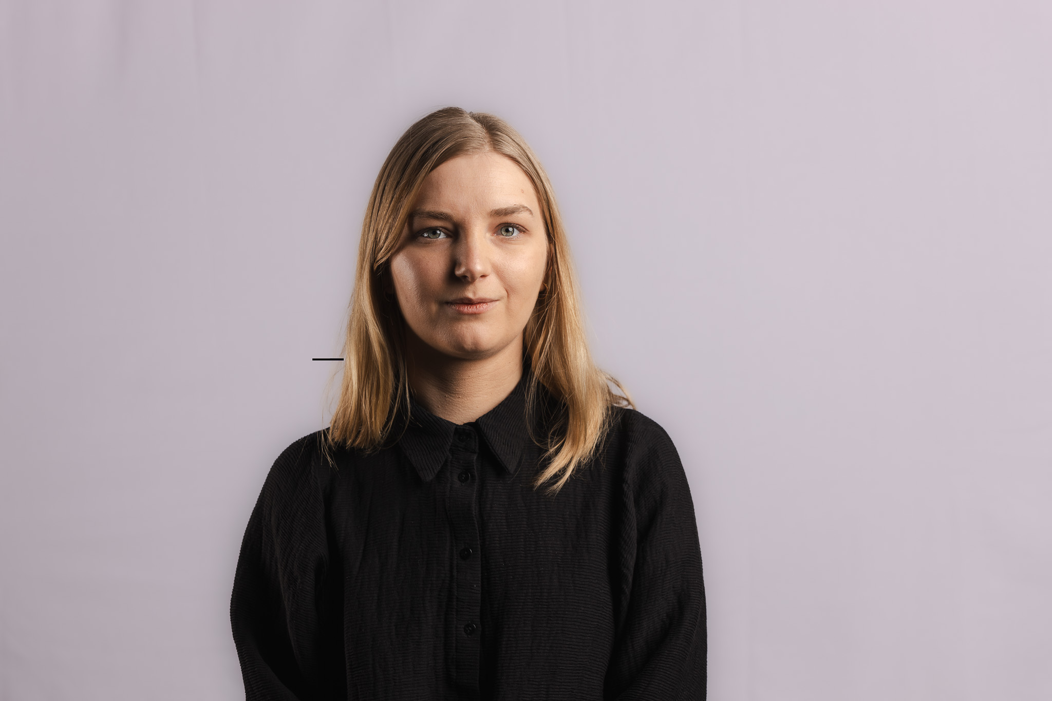 En kvinna med rakt blont hår och en svart skjorta står mot en ljus bakgrund och tittar in i kameran.