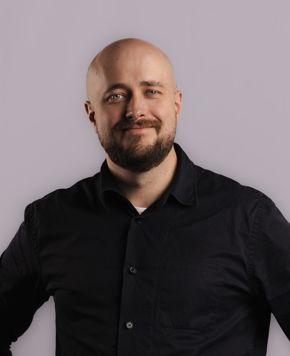 En man med skägg i mörk skjorta står mot en ljusgrå bakgrund, tittar in i kameran och ler svagt.