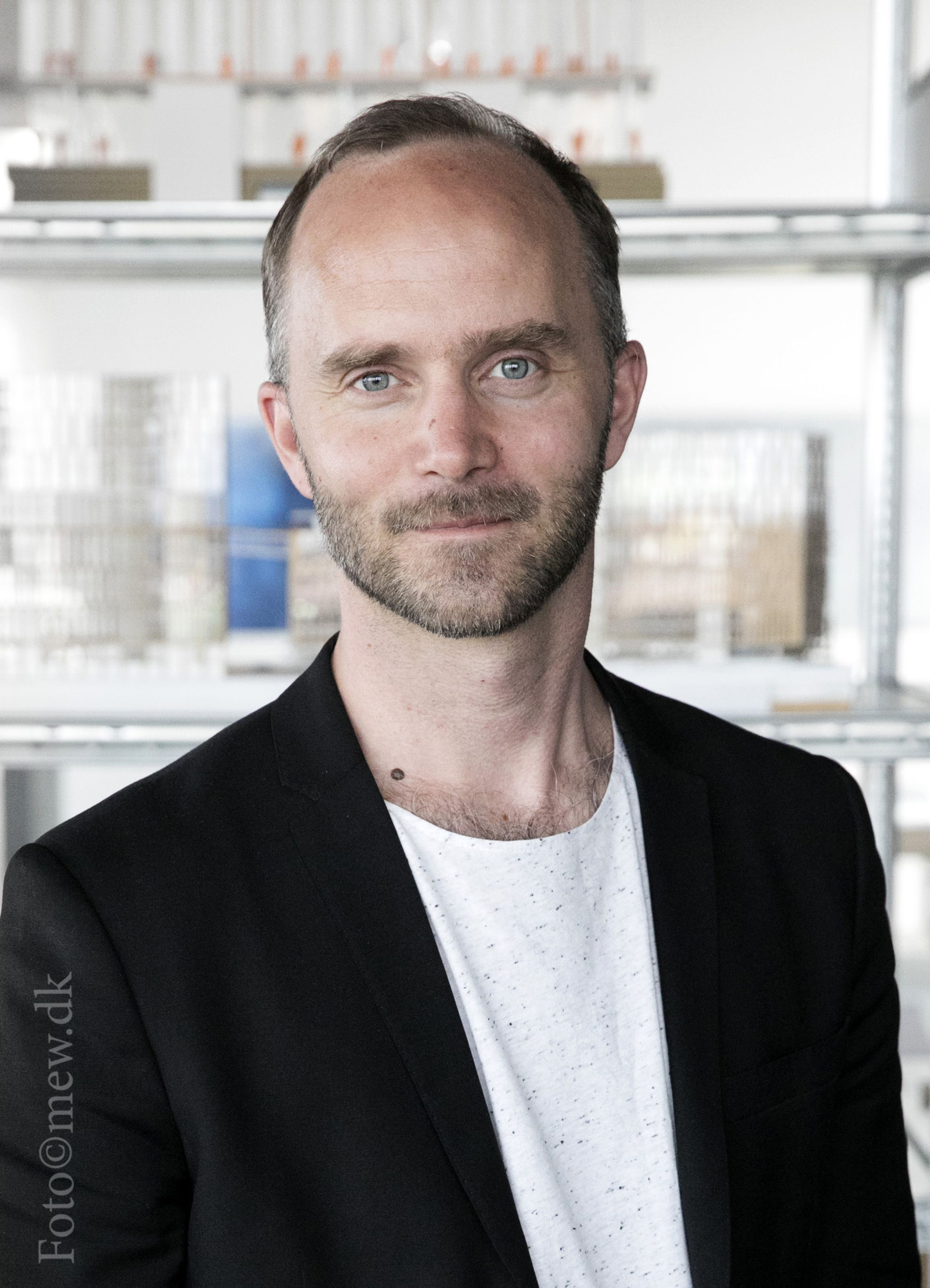 En man med kort hår och skägg, iklädd svart kavaj och vit skjorta, står inomhus framför hyllor med olika föremål.