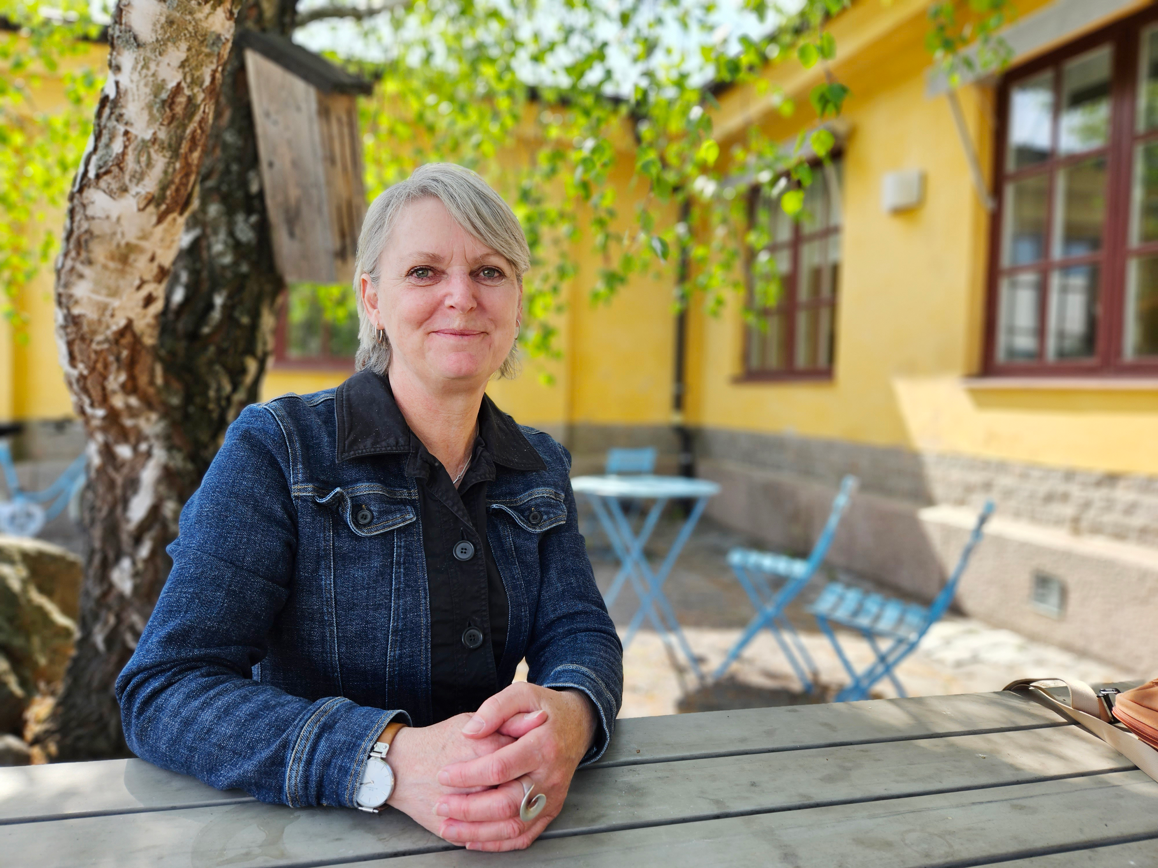 En kvinna med grått hår och jeansjacka sitter vid ett bord utomhus i en innergård med gula väggar och blå stolar.
