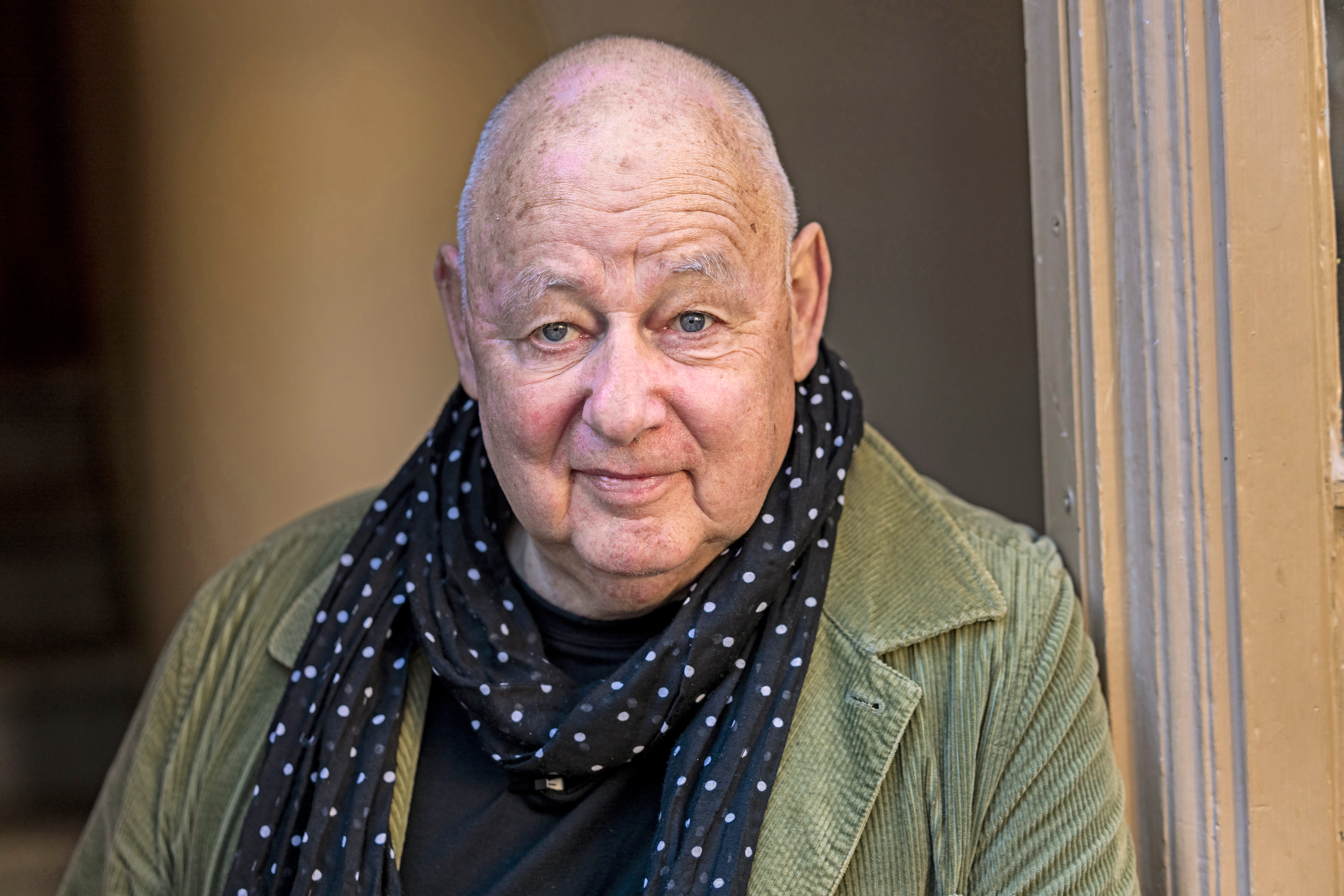 En äldre man med skalligt huvud, klädd i grön manchesterjacka, svart skjorta och svart scarf med vita prickar står inomhus vid en dörröppning av trä.