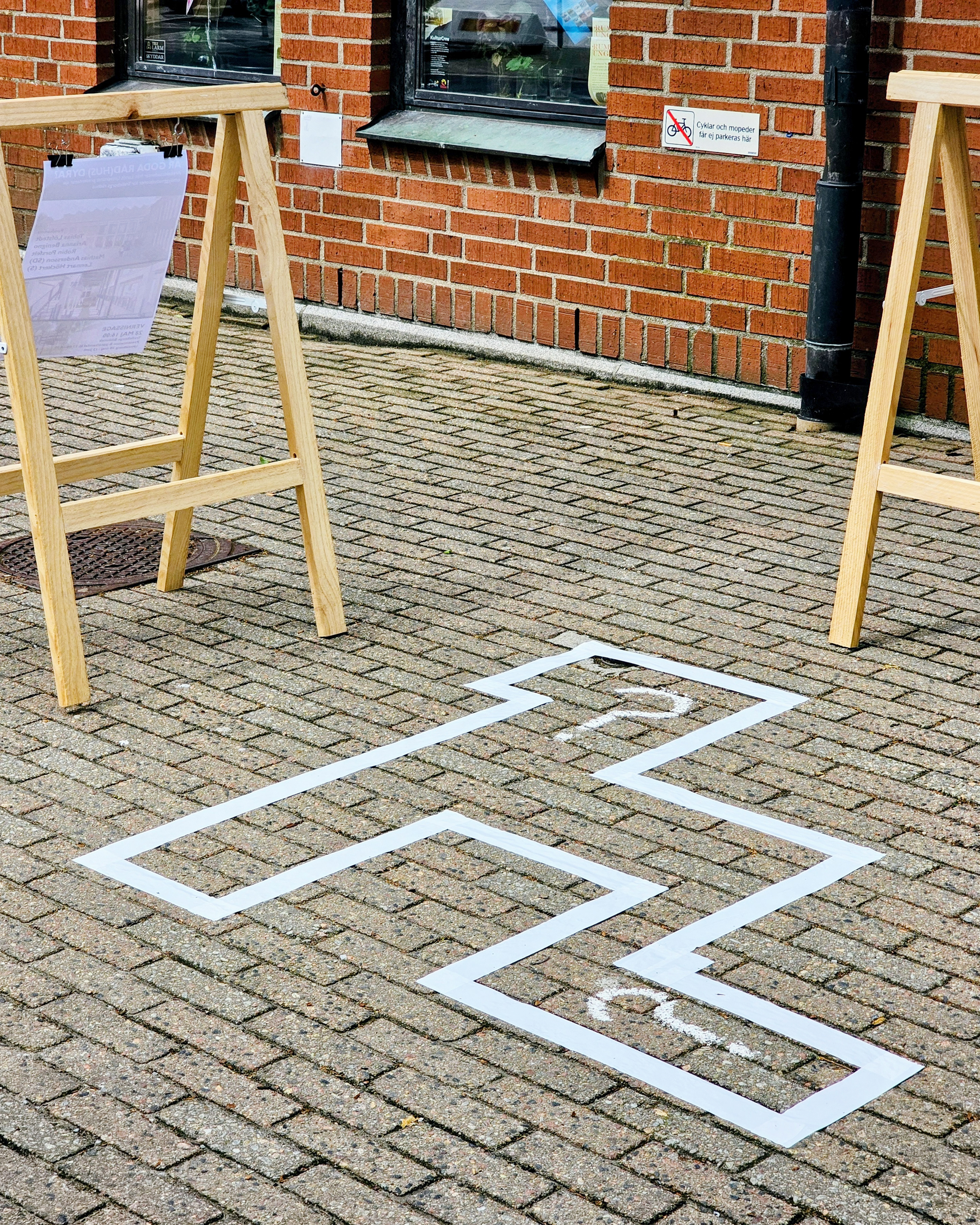 Vit tejp på ett tegelgolv markerar formen av en hopscotchbana mellan två trästafflier bredvid en tegelbyggnad.