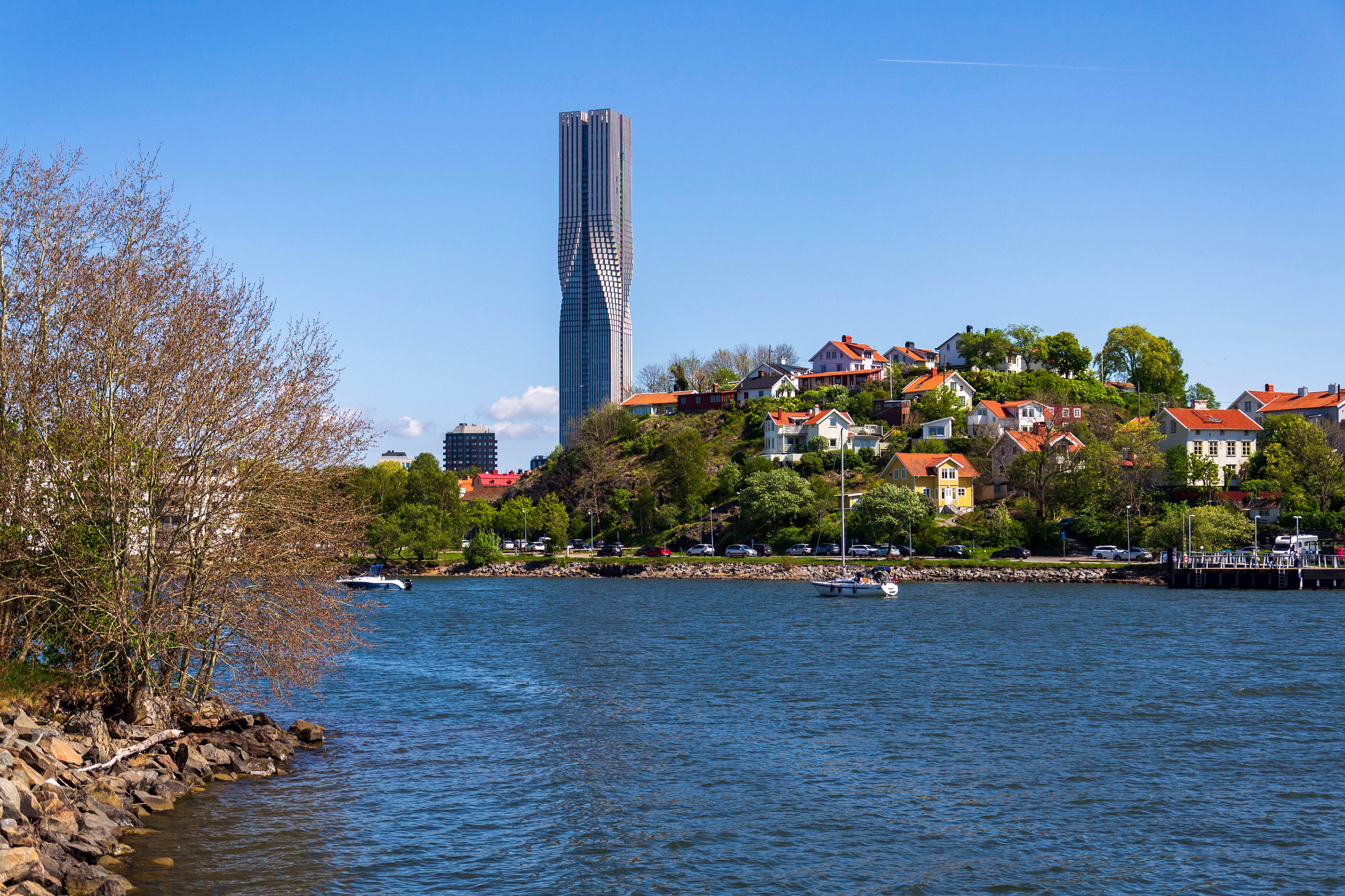 En hög skyskrapa står bakom en sluttning med hus och växtlighet, intill ett vattendrag där det finns båtar.