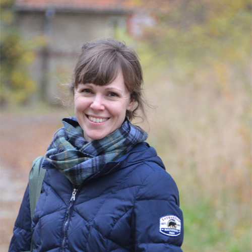 En kvinna med blå jacka och rutigt skärp står utomhus och ler mot kameran. I bakgrunden finns växter och en byggnad.