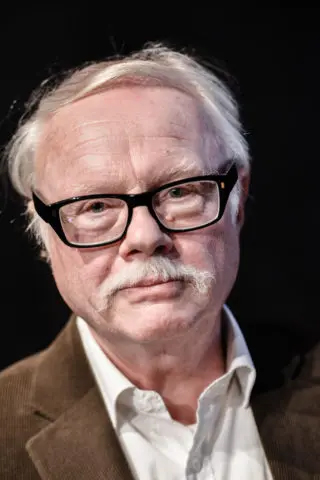 En äldre man med ljus hy, vitt hår, mustasch och svarta glasögon har brun jacka och vit skjorta, tittar mot kameran.