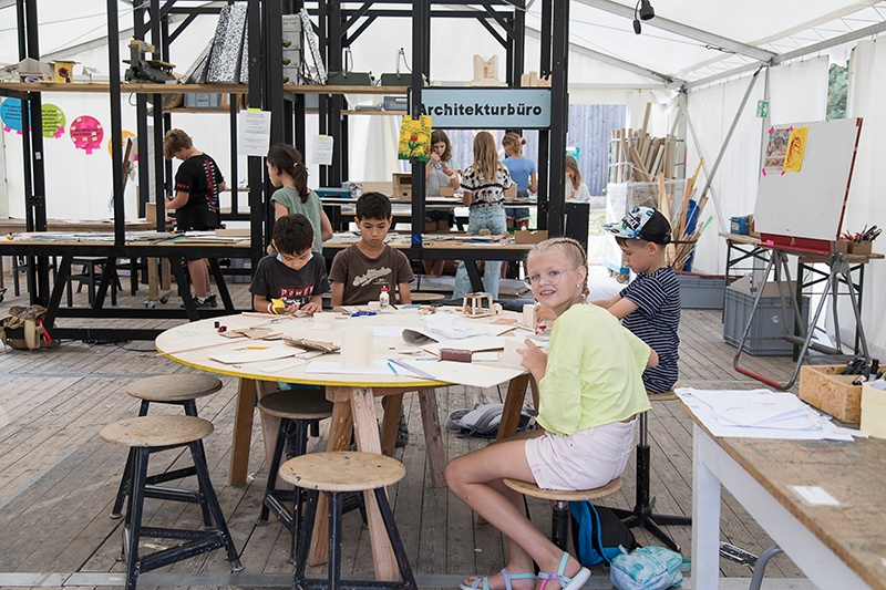 Barn sitter runt ett bord och arbetar med pyssel i en verkstad med skylten "Architekturbüro", andra barn och material syns i bakgrunden.