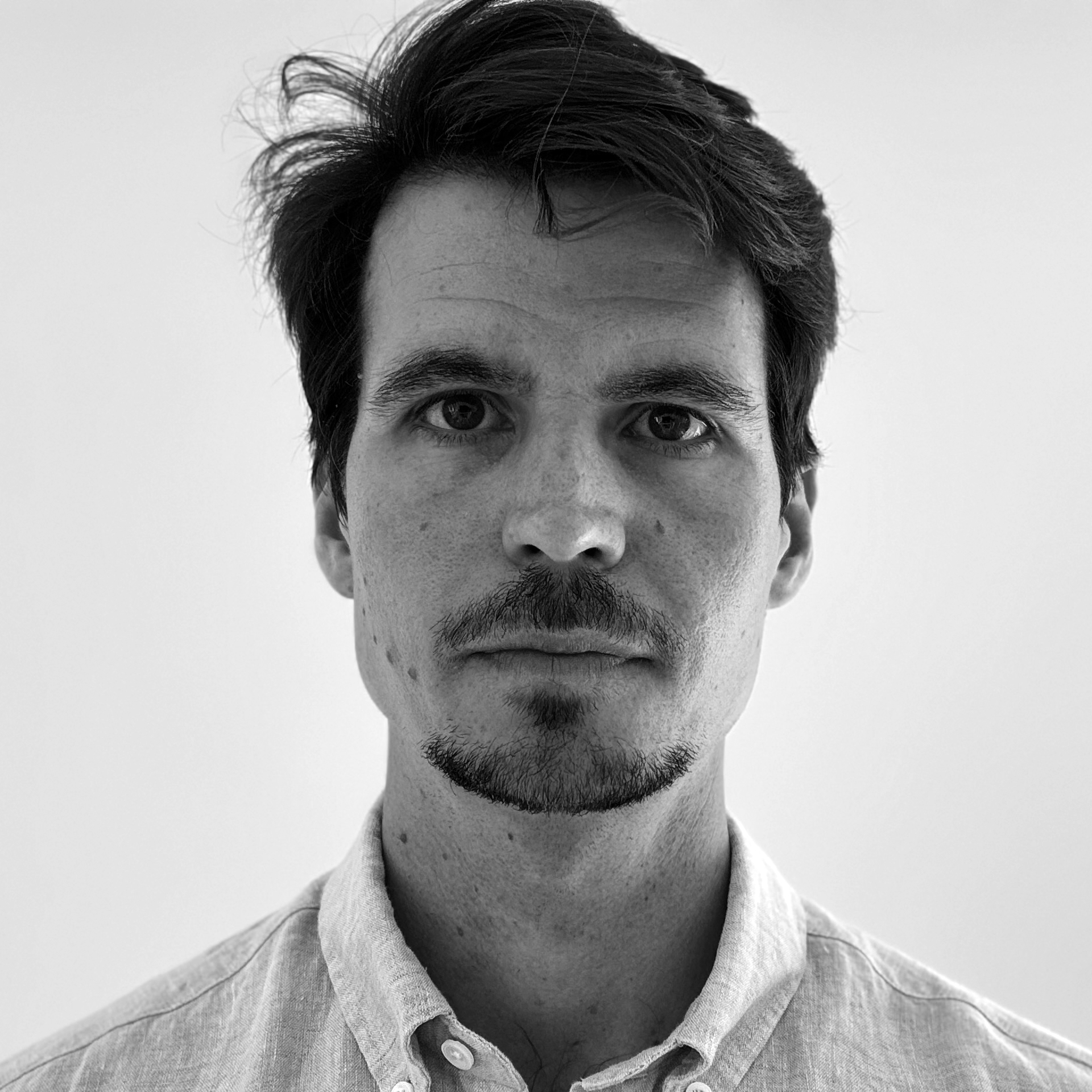 En man med kort mörkt hår och lite skägg står vänd mot kameran framför en ljus bakgrund.