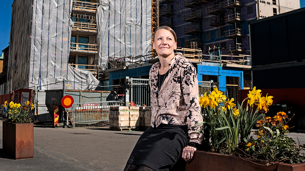 En kvinna sitter på en planteringslåda med gula blommor framför en byggarbetsplats med byggnadsställningar och halvfärdiga hus.
