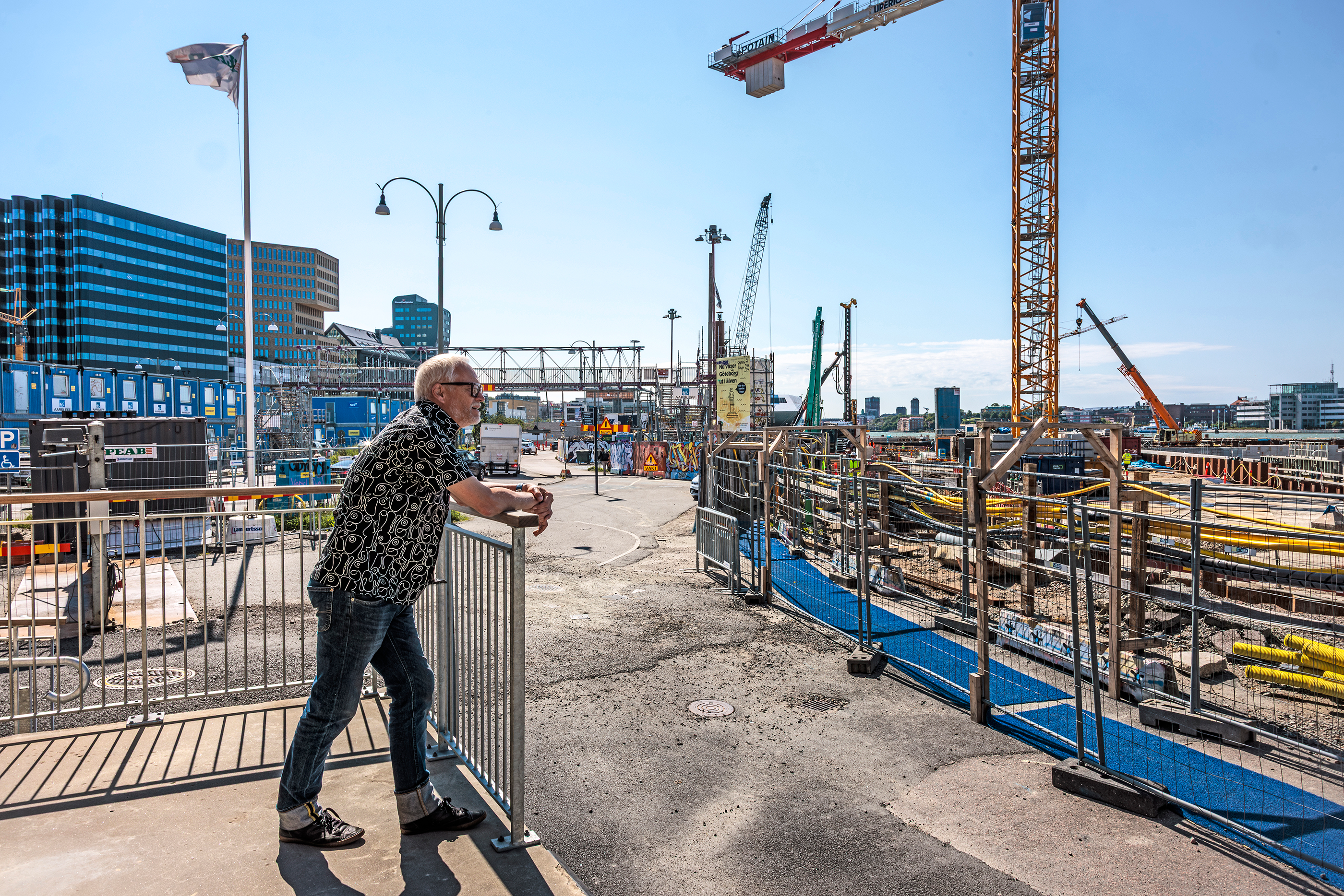 En äldre man lutar sig mot ett räcke och tittar på en byggarbetsplats med kranar och byggmaterial under dagen.