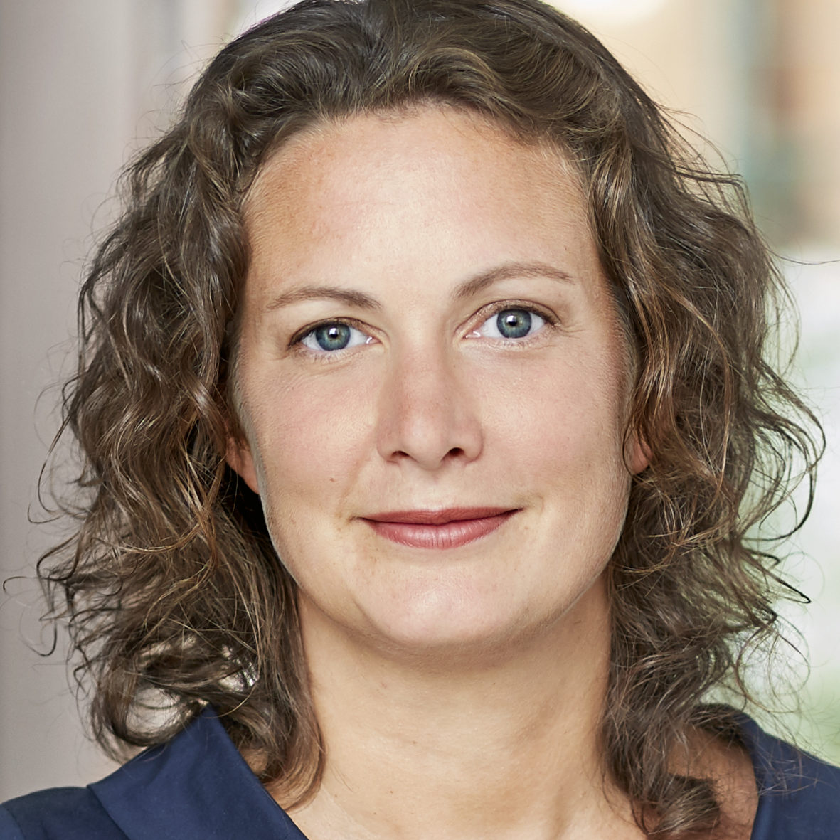 En kvinna med brunt, vågigt hår och blå ögon, iförd en marinblå topp, ser mot kameran och ler svagt. Bakgrunden är suddig.